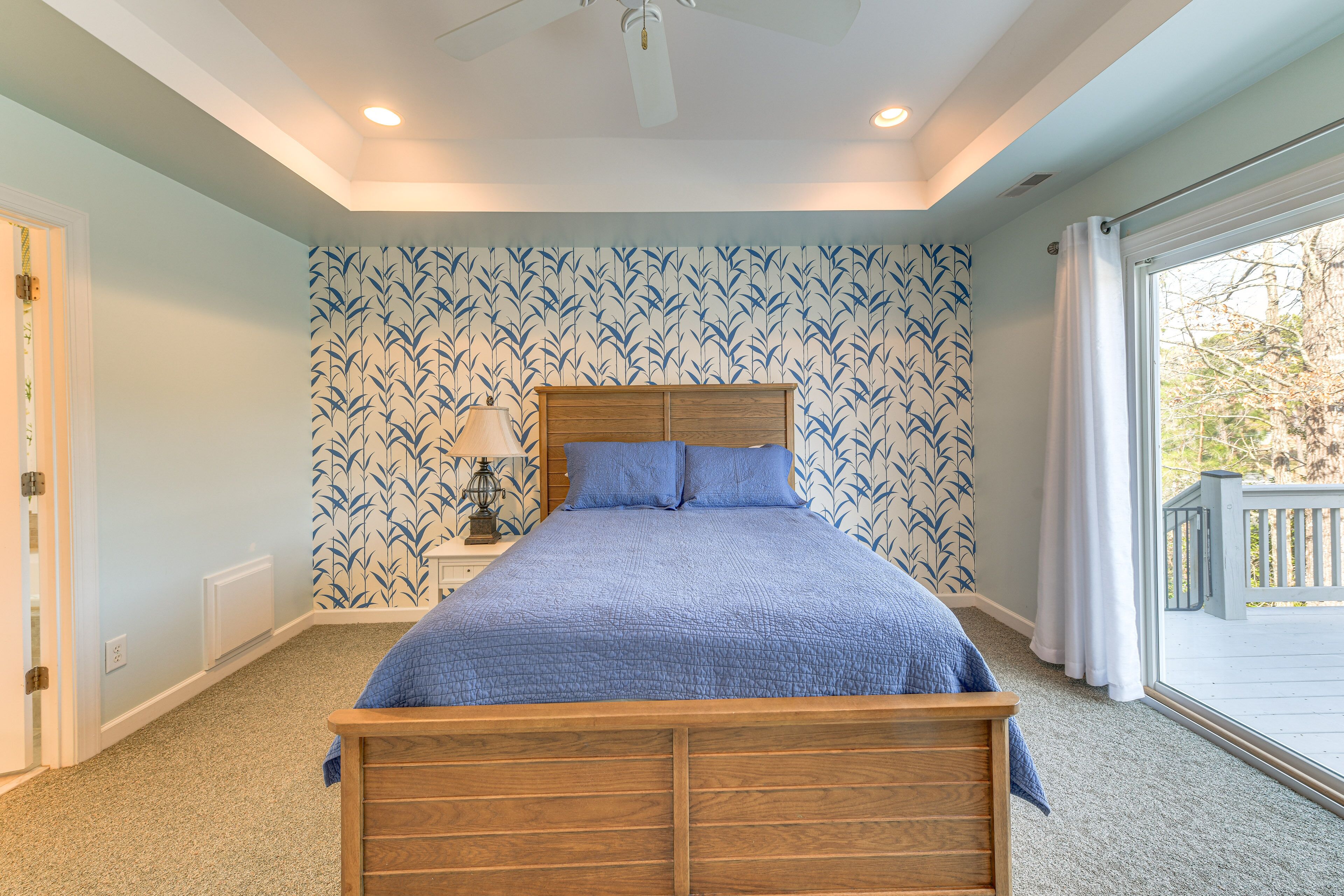 A bedroom with a wooden bed, a nightstand with a lamp, a window with a view of the outside, and a patterned wallpaper.