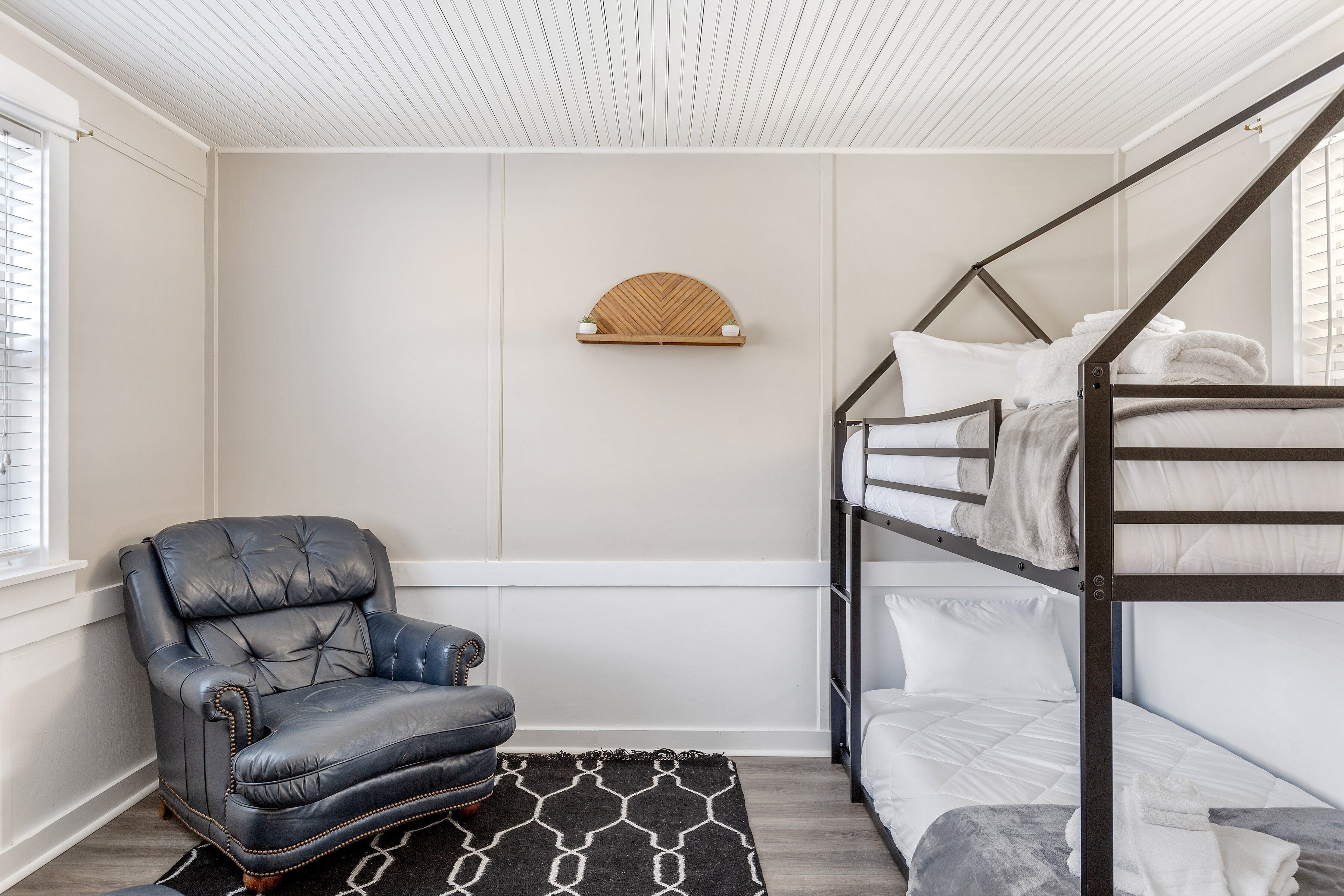 A bunk bed with white bedding, a leather armchair, and a patterned rug in a room with white walls and a wooden ceiling.