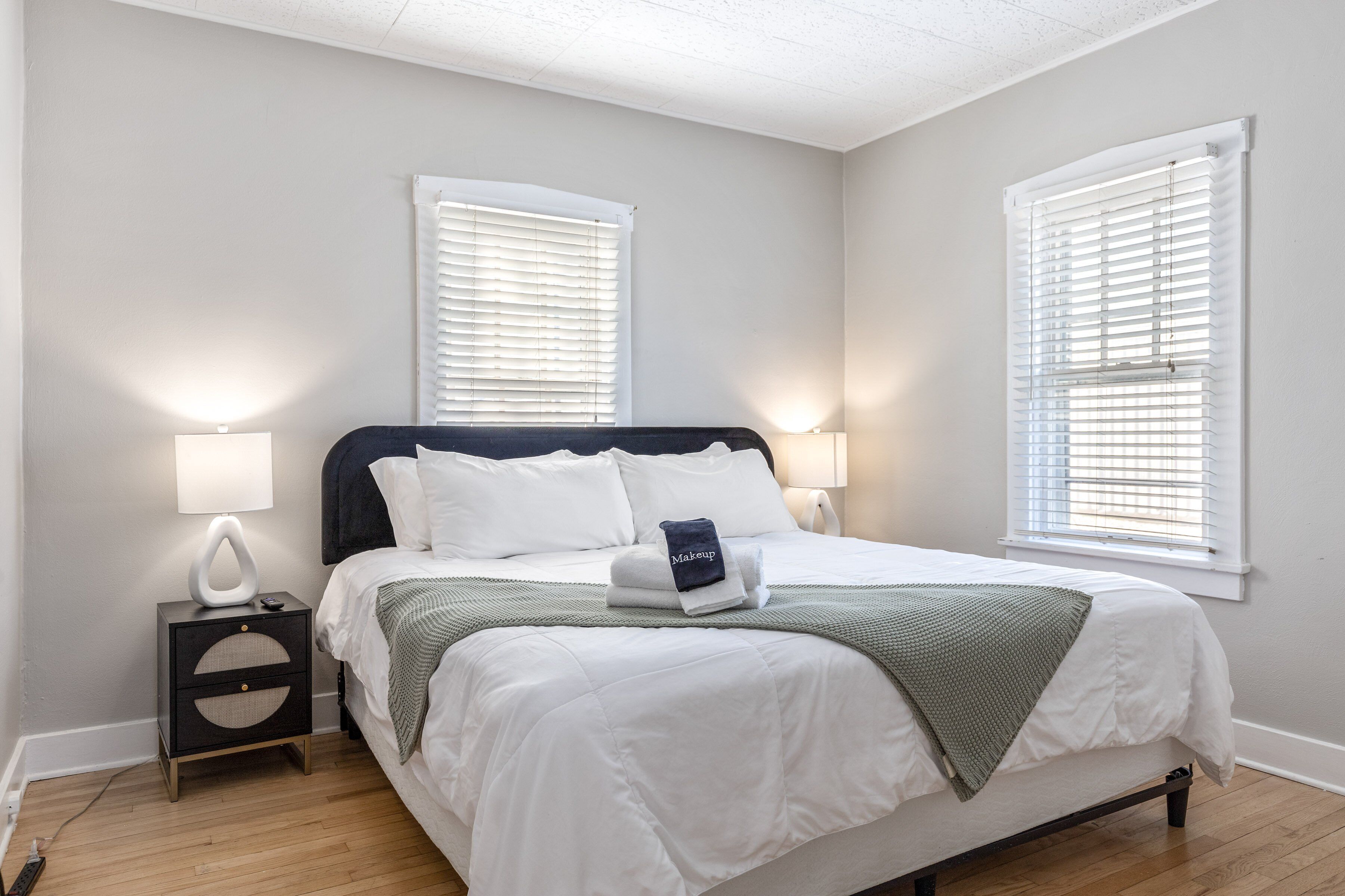 A neatly made bed with white bedding and a green throw blanket, flanked by two bedside lamps and a small nightstand with a lamp.