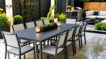 An outdoor patio with a rectangular table and chairs, surrounded by potted plants and a grill.