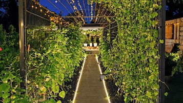 A pathway flanked by a climbing vine structure with lights, leading to a seating area.