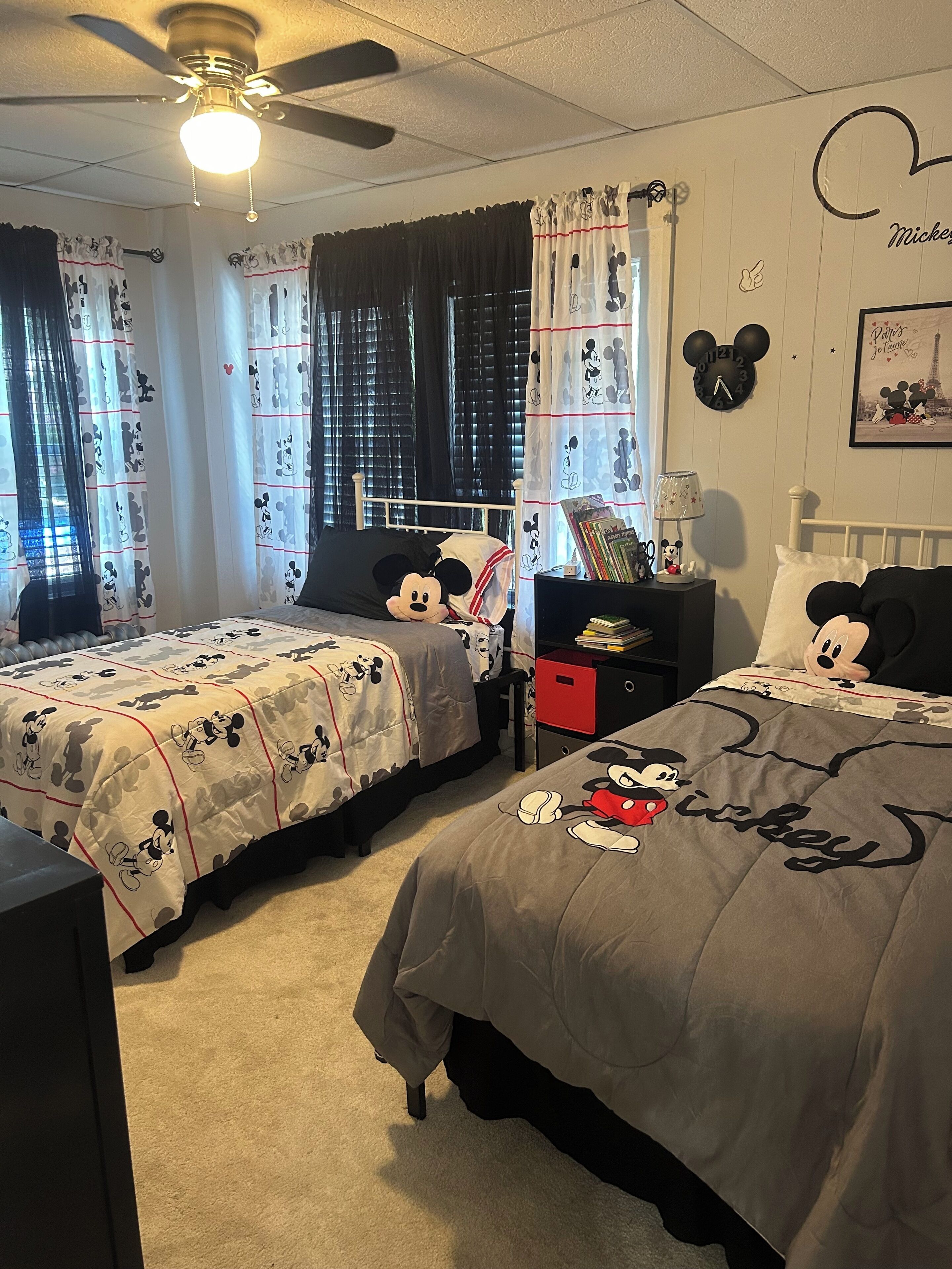 Une chambre avec deux lits, chacun agrémenté de linge de lit aux motifs de Mickey Mouse. Sur l’un des lits, on trouve un oreiller à l’effigie de Mickey Mouse, sur une table de chevet, une lampe Mickey Mouse, et sur le mur, une horloge Mickey Mouse.