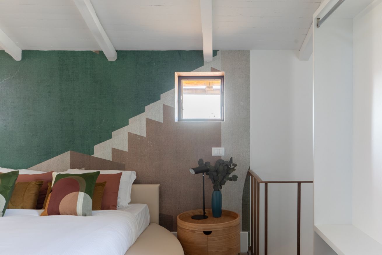 A bedroom with a bed, pillows, a nightstand, a window, and a staircase.