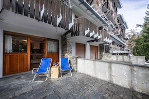 Un hôtel doté d’une façade en pierre, de balcons en bois et d’un espace extérieur aménagé avec une terrasse pavée, deux fauteuils bleus et des verres à vin.