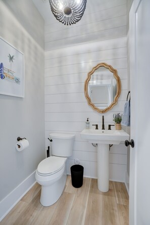 Et badeværelse med et hvidt toilet, en håndvask på fod, et spejl med træramme, et håndklædestativ og et billede i ramme på væggen.