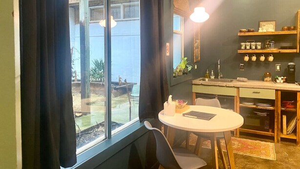 A compact kitchen with a dining area, a large window with curtains, and a view of an outdoor area. - Downtown Lynchburg, Vintage Courtyard Loft, Va (Lynchburg)