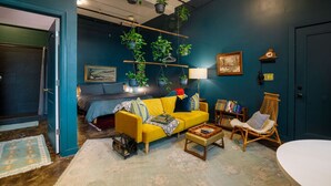 A cozy living room with a yellow sofa, a wooden coffee table, and a bookshelf. There are hanging plants, a lamp, and a painting on the wall. - Downtown Lynchburg, Vintage Courtyard Loft, Va (Lynchburg)