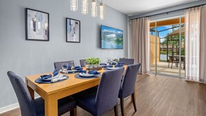 Zona pranzo con tavolo di legno apparecchiato per sei persone, sedie blu e un'ampia finestra con vista su giardino e edificio.