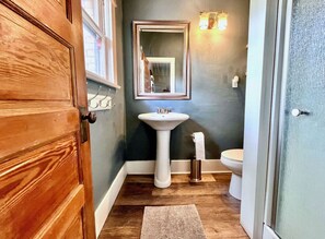 Un baño con puerta de madera, lavamanos pedestal, inodoro y espejo con luces empotradas.