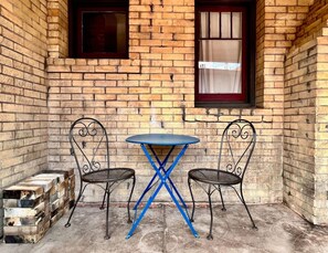 Un pequeño espacio al aire libre con una mesa redonda y dos sillas, frente a un muro de ladrillo con ventanas.
