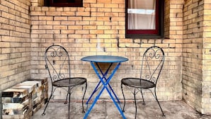 Un pequeño espacio al aire libre con una mesa redonda y dos sillas, frente a un muro de ladrillo con ventanas.