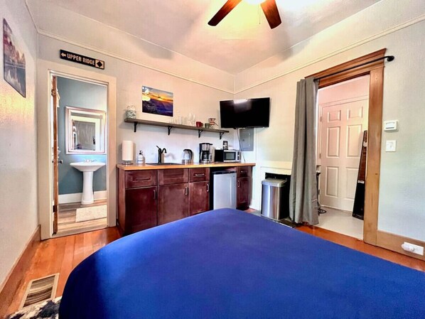 Un dormitorio con una cama grande, un televisor montado en la pared, una zona de cocina con microondas, lavabo y armarios, y una puerta con la leyenda 'Upper Ridge'.