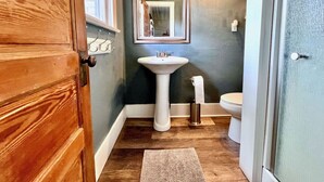 Un baño con puerta de madera, lavamanos pedestal, inodoro y espejo con luces empotradas.