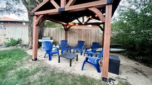 A wooden pergola structure with blue chairs and a table, surrounded by a grassy yard and a wooden fence. - Relaxing Historic Home in Downtown Pet Friendly (Flagstaff)