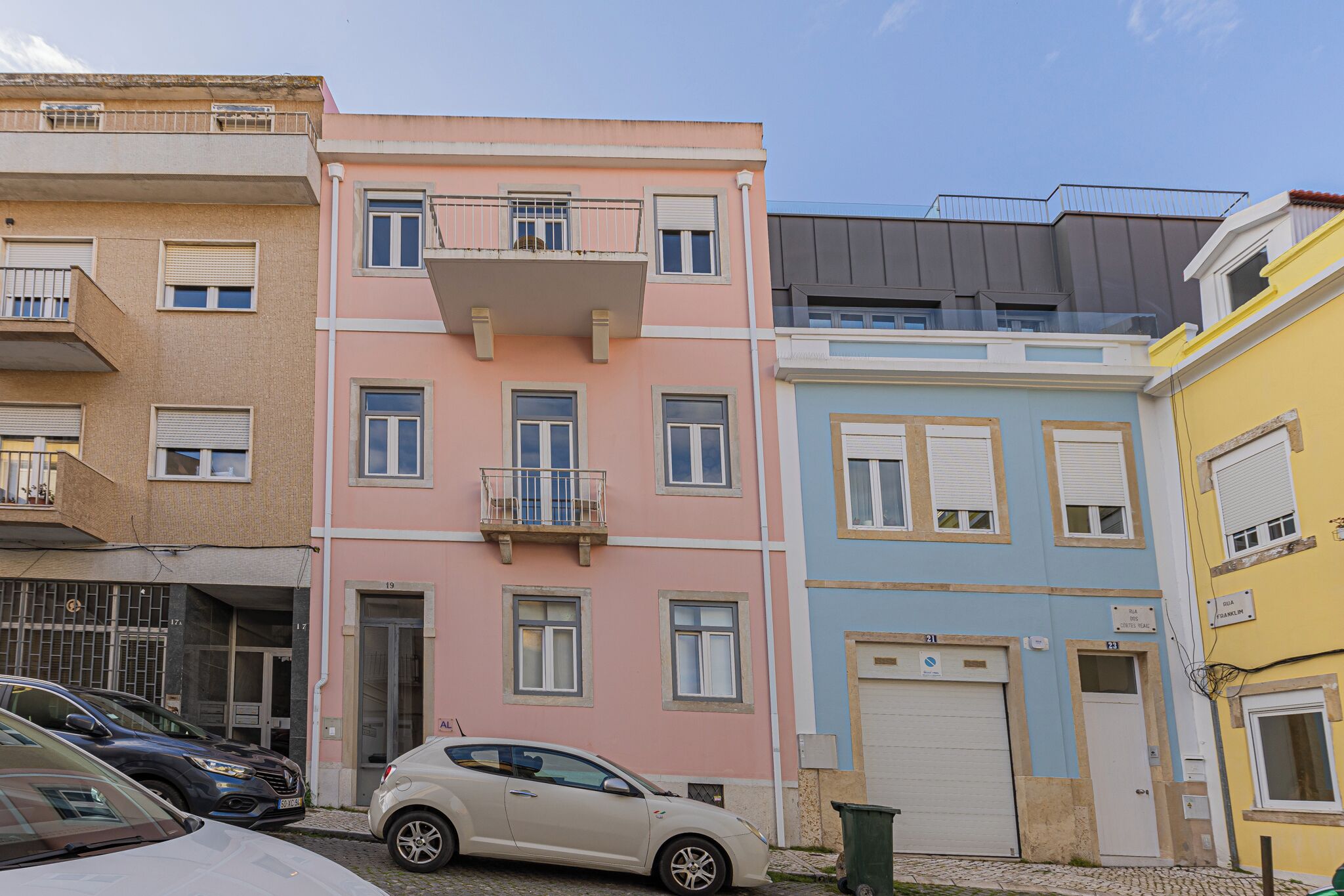 A row of multi-story buildings with balconies, a parking area, and a clear sky.