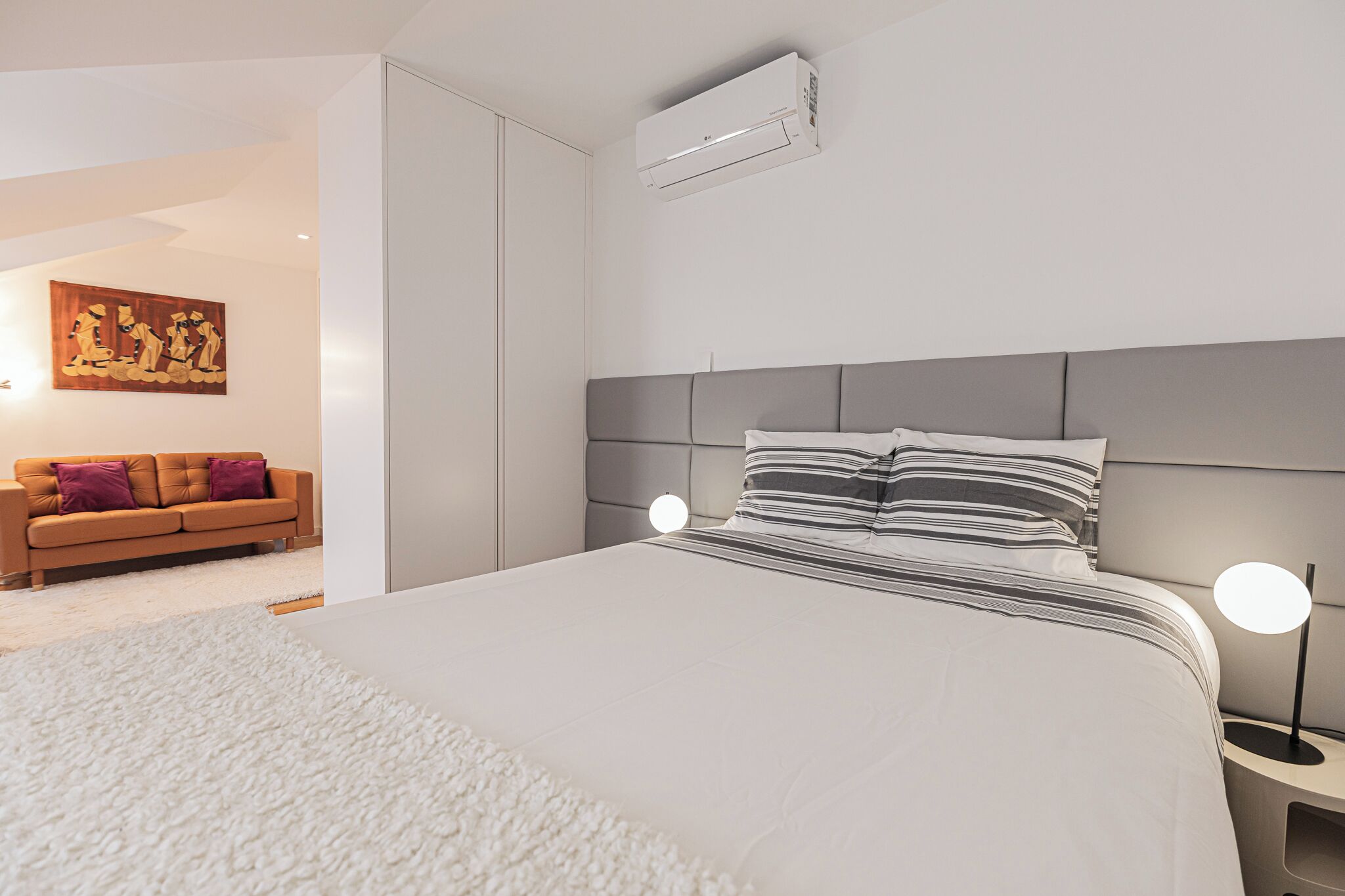 A modern bedroom with a large bed, a bedside table with a lamp, a sofa in the background, and an air conditioning unit on the wall.