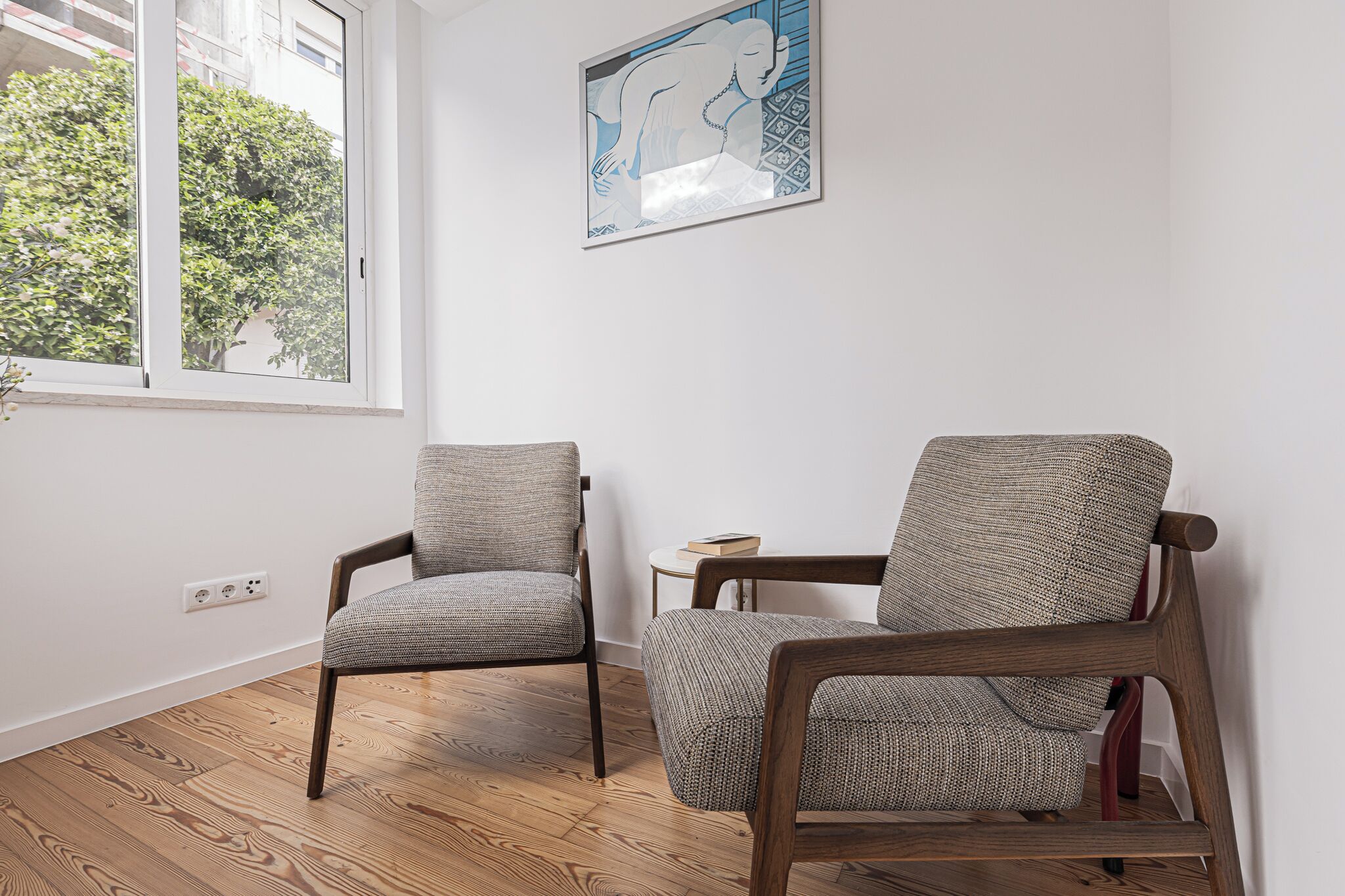 A room with two chairs, a small table, a painting on the wall, and a window with a view of trees.