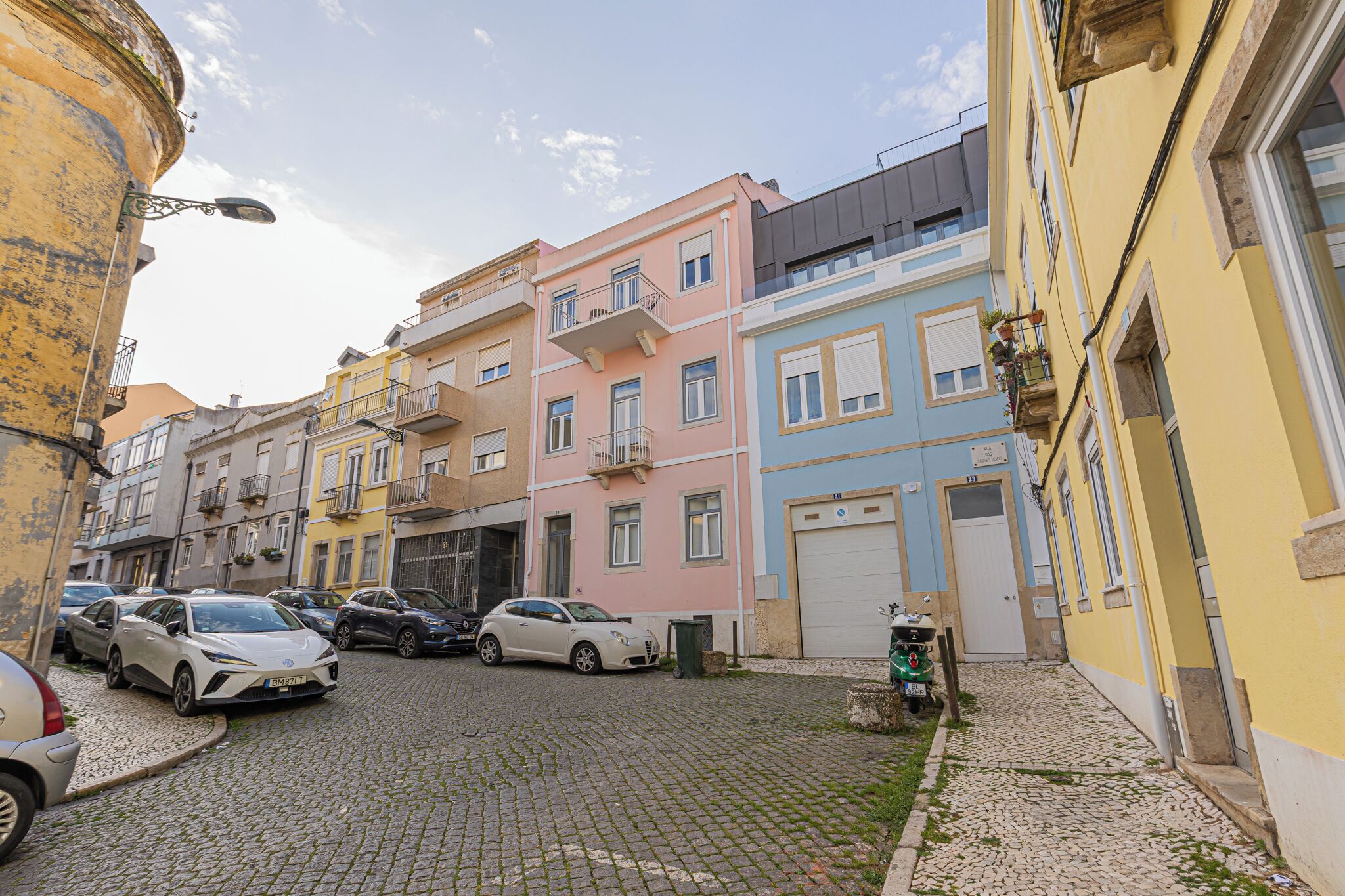 A cobblestone street lined with colorful buildings, parked cars, and a green motorcycle.