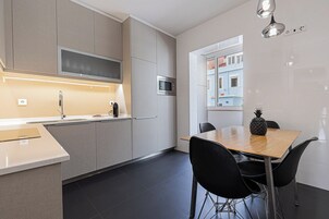Uma cozinha moderna com micro-ondas embutido, uma mesa de jantar de madeira com cadeiras pretas e vista para um edifício azul pela janela.