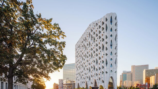 A modern building with a unique geometric facade, surrounded by a city street with pedestrians and vehicles.