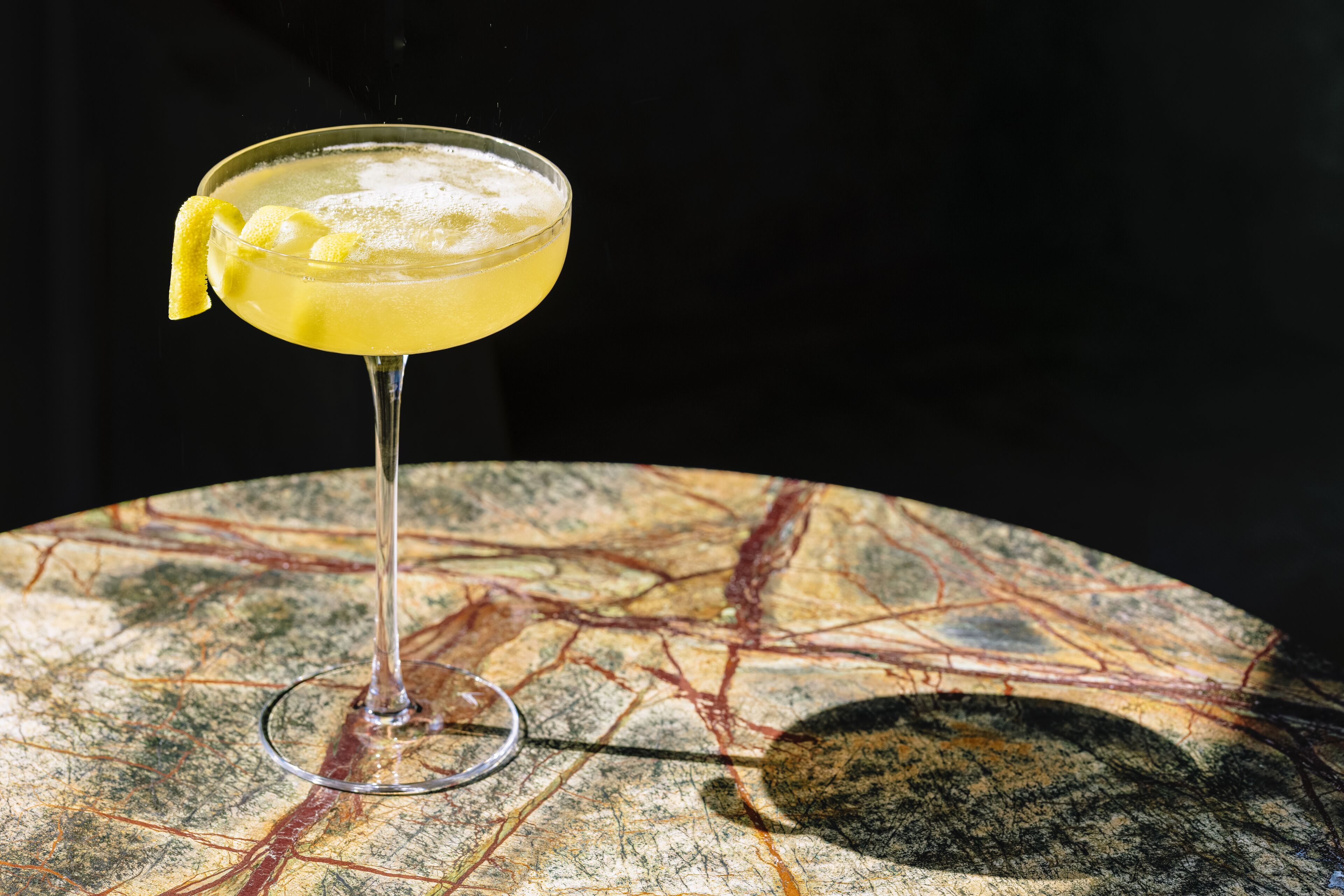 A cocktail in a glass with a lemon peel garnish on a table with a marbled surface.