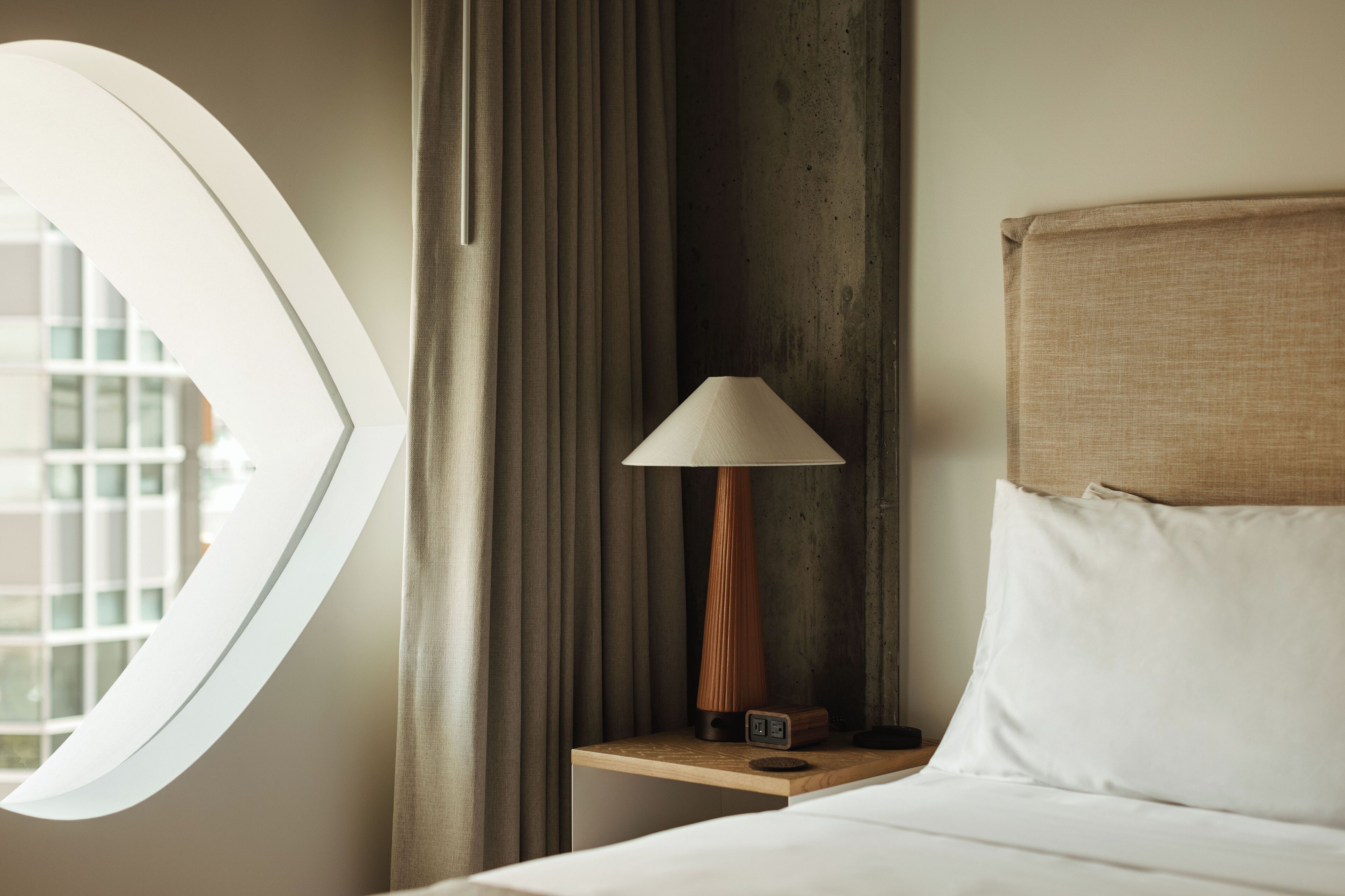 A hotel room with a bed, a nightstand with a lamp, a window with curtains, and a view of buildings outside.