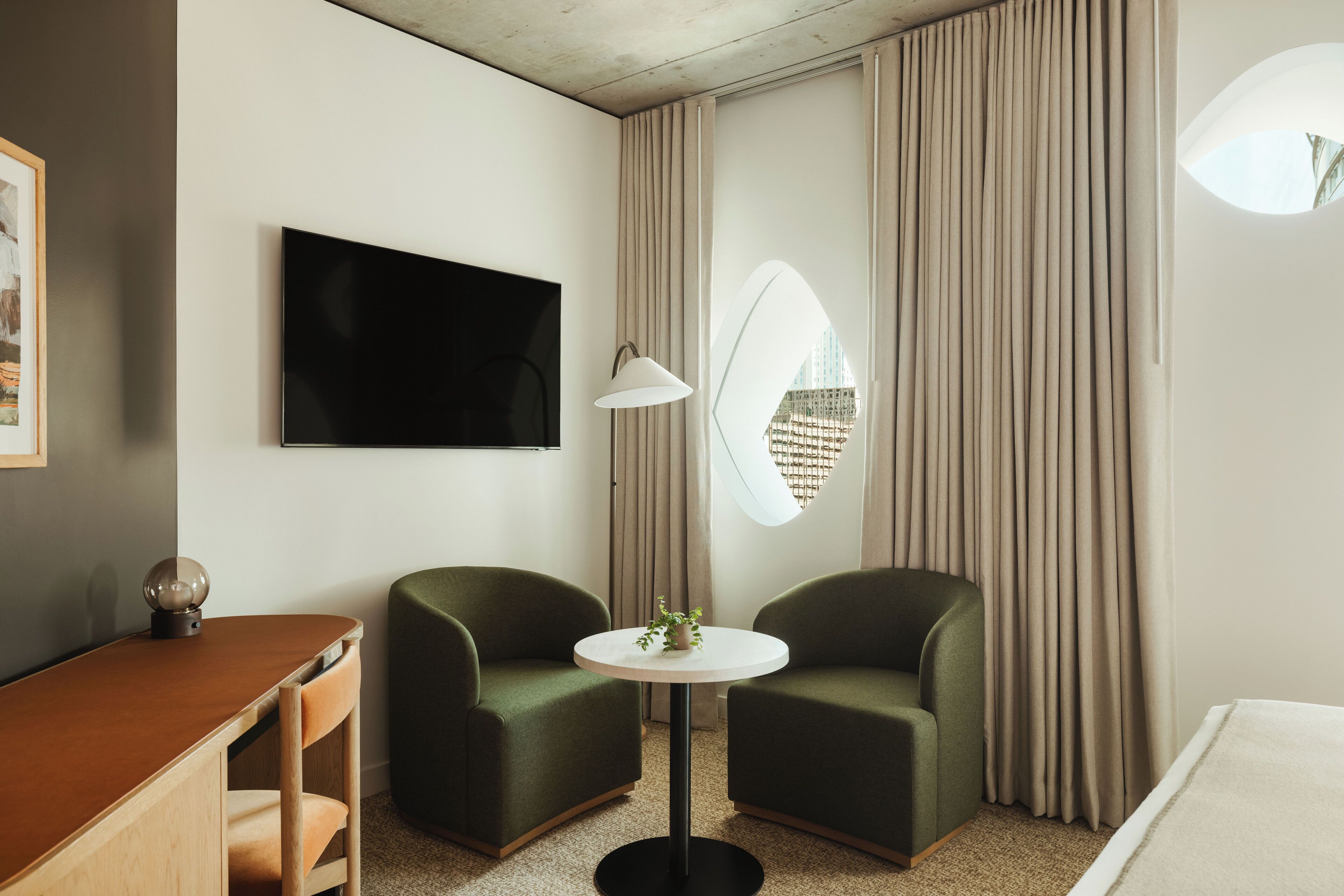 Ein modernes Hotelzimmer mit Flachbildfernseher, einem runden Tisch mit Pflanze, zwei grünen Sesseln, einem Schreibtisch und einem großen Fenster mit Vorhängen.