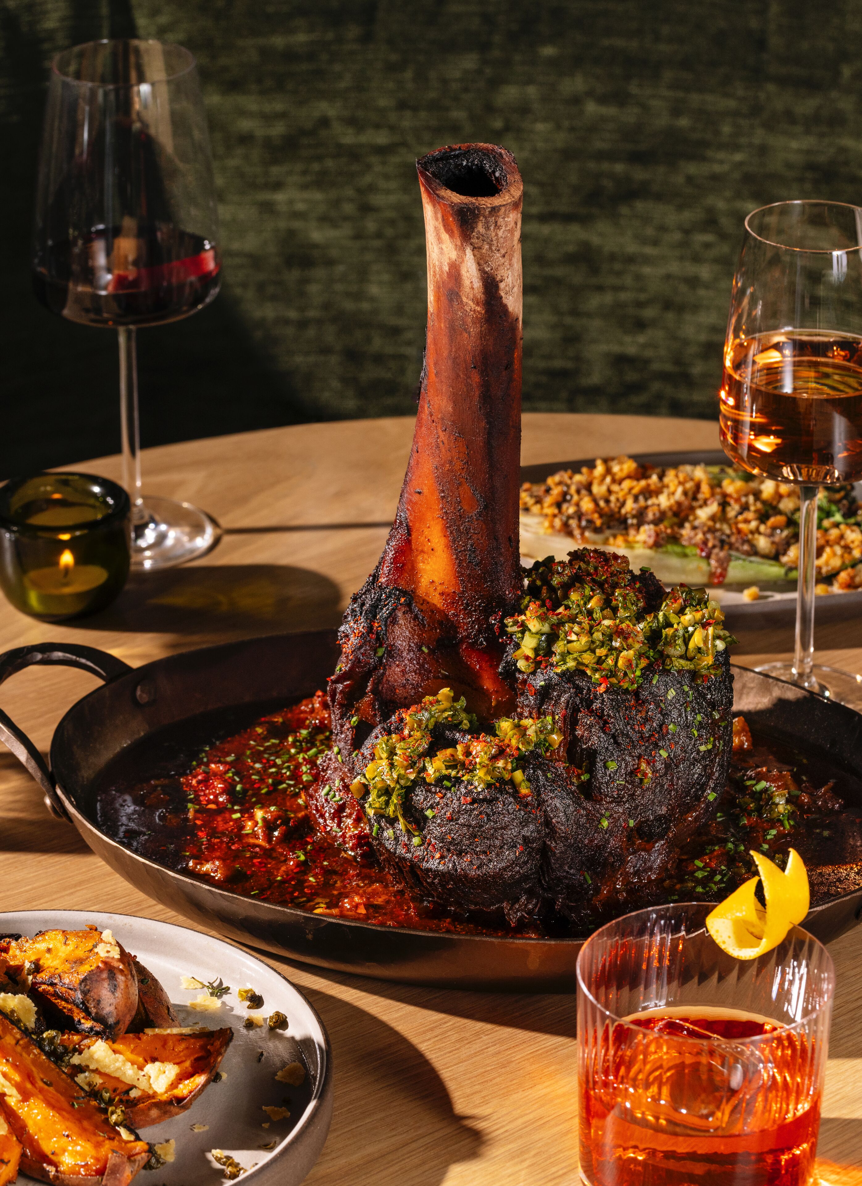 A dining table with a roasted leg of lamb, a pan of food, and glasses of wine.