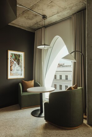 A modern room with a round table, two green armchairs, a framed painting, and a view of buildings through large windows.