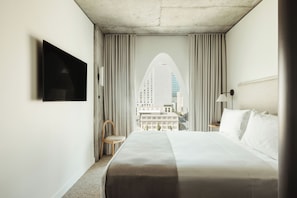 Habitación de hotel con una cama grande, un televisor montado en la pared, una silla y vistas de un paisaje urbano a través de una ventana con cortinas.