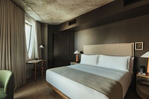 A hotel room with a large bed, a desk, a chair, a wardrobe, and a nightstand with a lamp.
