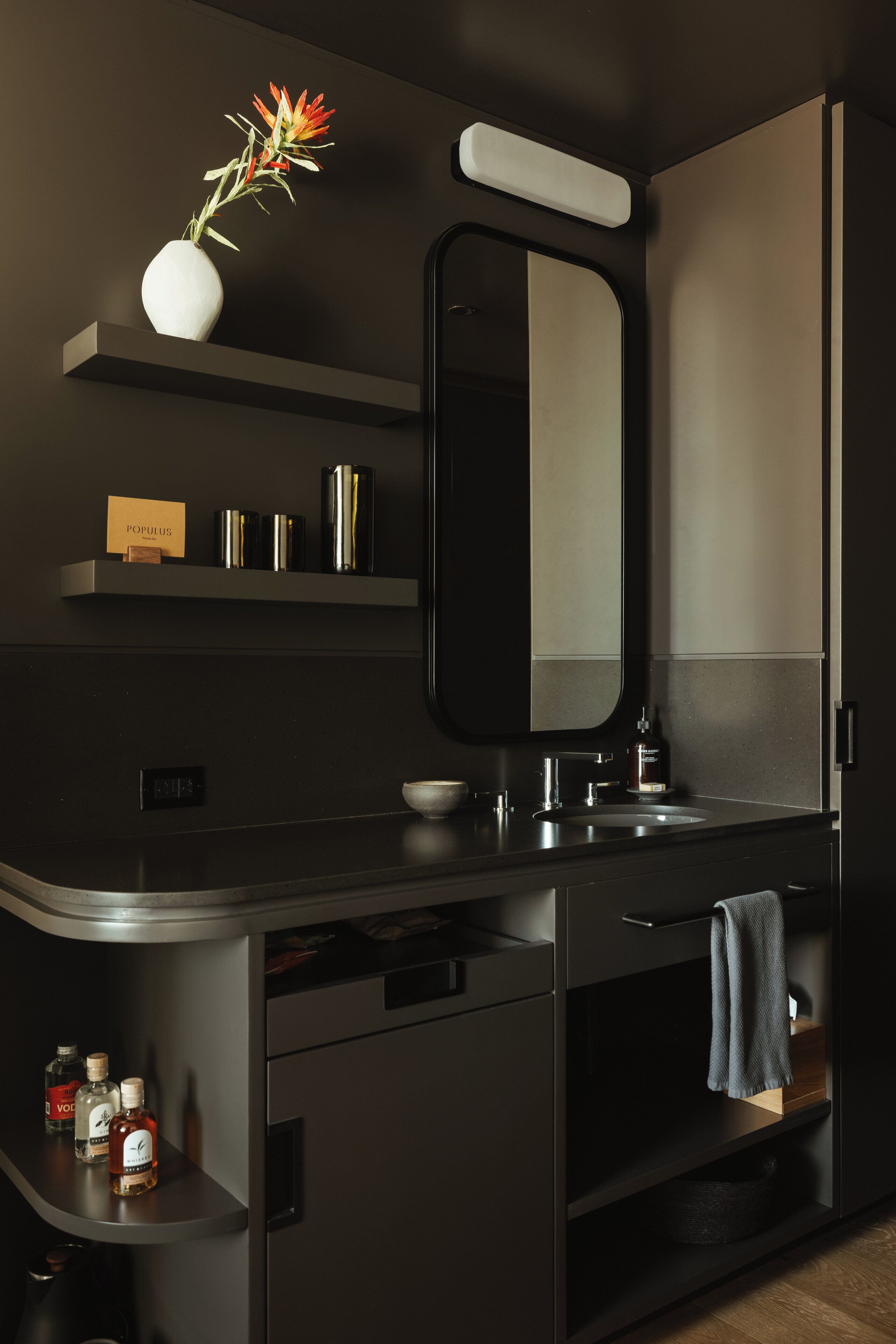 Une salle de bain moderne aux couleurs sombres, avec un miroir, un lavabo et une étagère contenant des articles de toilette.