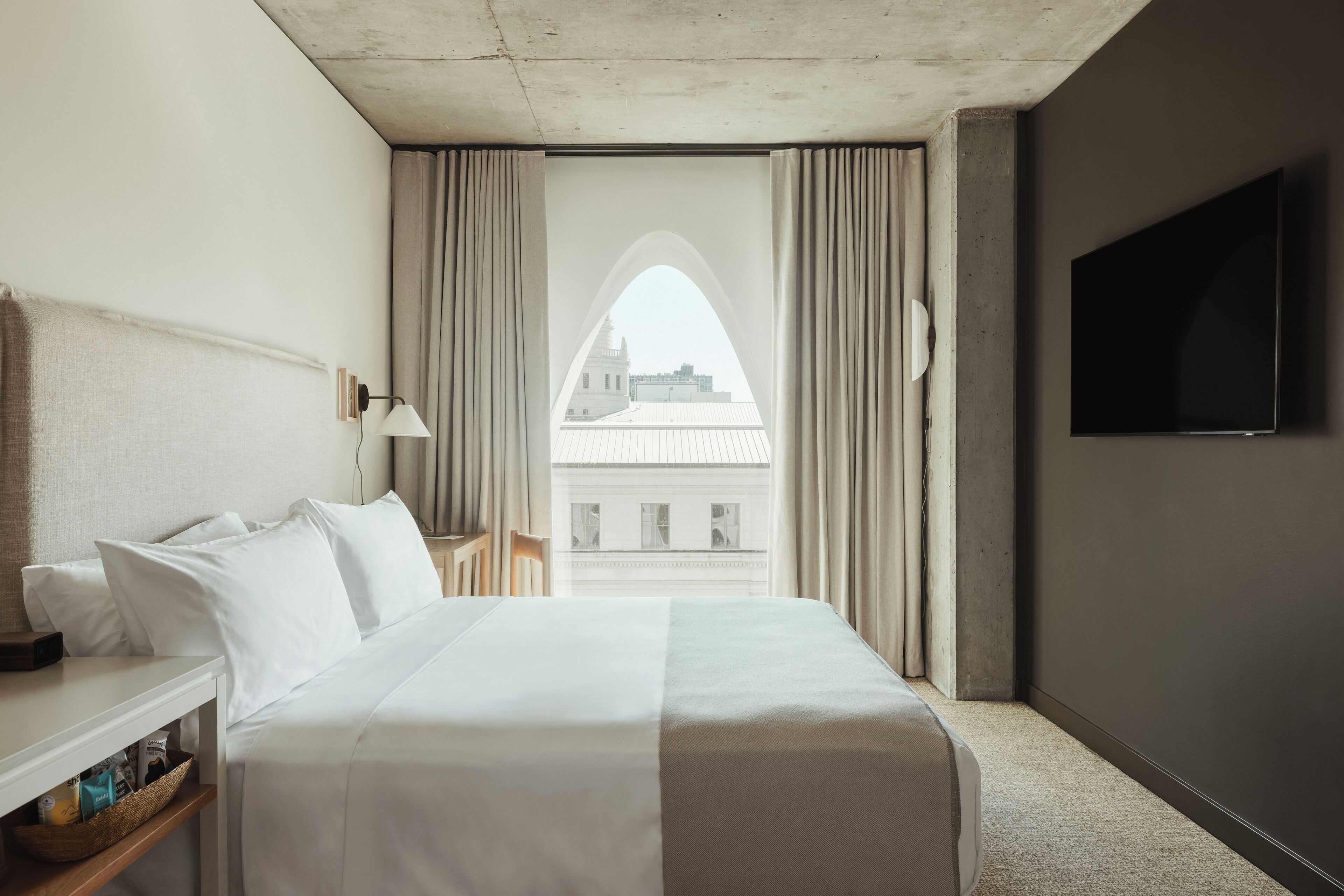 Hotelkamer met een bed, nachtkastje, lamp, televisie en uitzicht op een gebouw door een raam met gordijnen.