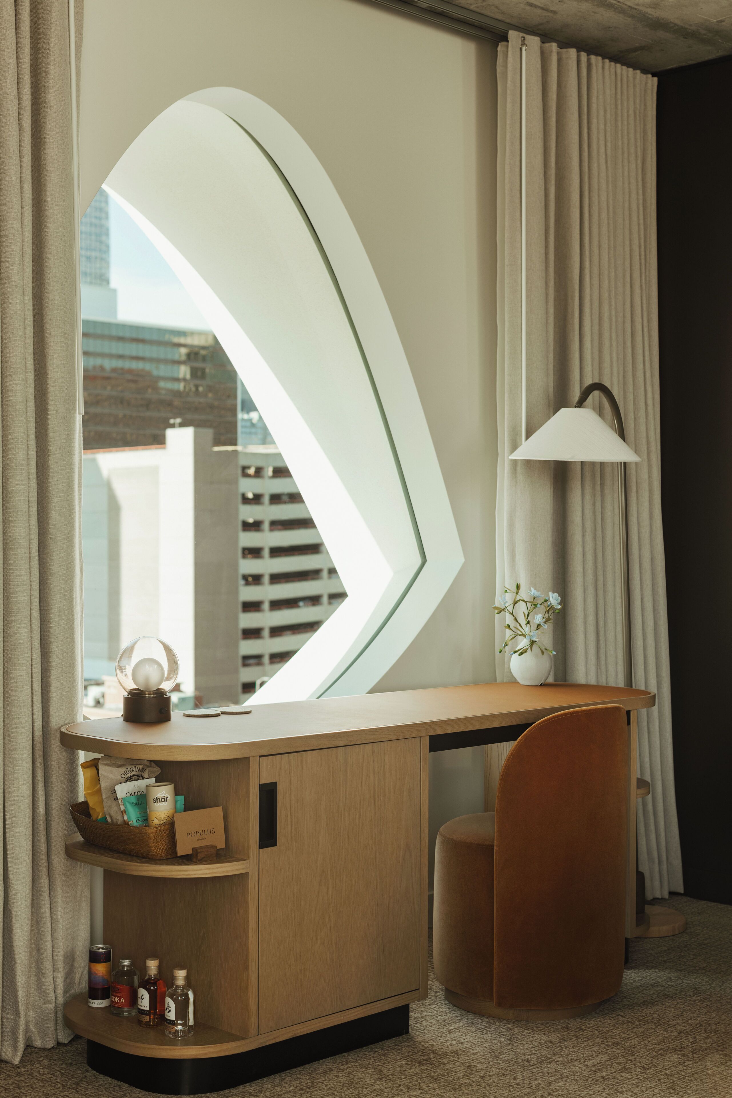 A modern hotel room with a large window, a wooden desk, a small chair, and a lamp.