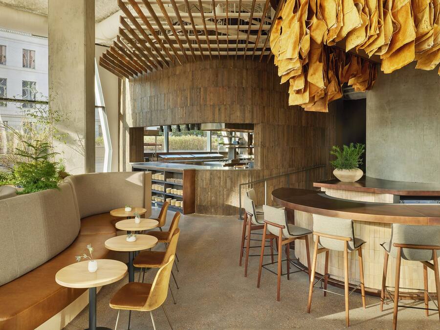 Uma área de bar moderna com balcão curvo, banquetas de madeira e um espaço de estar com mesas redondas e bancos acolchoados.