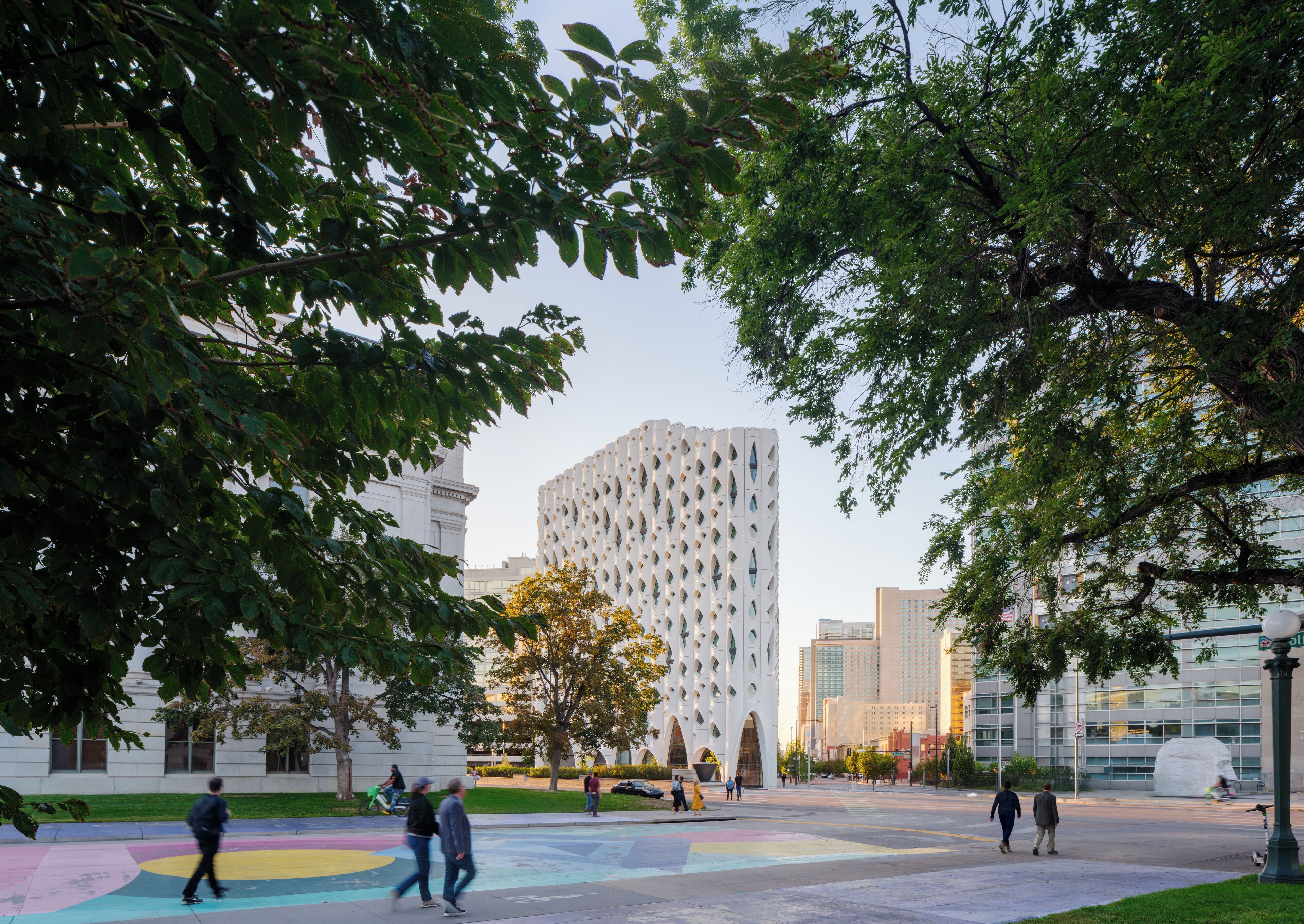 Ein modernes Gebäude mit einer markanten Fassade, umgeben von Bäumen und einem öffentlichen Platz mit Passanten.