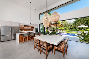 Une cuisine moderne dotée d’un grand îlot central, d’appareils électroménagers en acier inoxydable et d’un coin repas offrant une vue sur une piscine et un jardin.