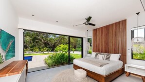 A modern bedroom with a large bed, a ceiling fan, a wooden slatted wall, and a view of a garden through a sliding glass door. - Beachside Bliss 4BR Lux Villa, Pool, Terrace, Golf (Pinilla)