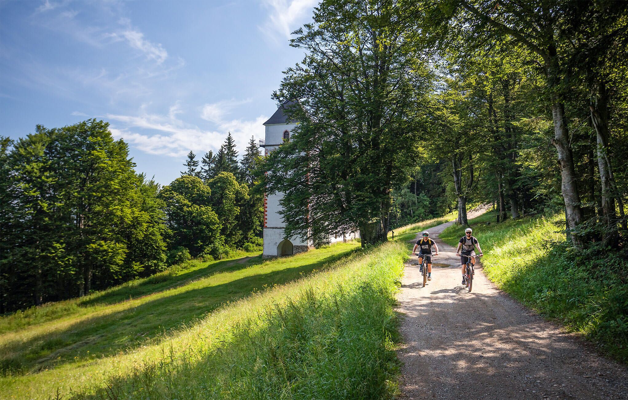 Un chemin de terre où l’on voit des cyclistes, entouré d’une végétation luxuriante et bordé d’un bâtiment doté d’une tour.