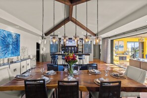 A spacious dining area with a wooden table set for six, surrounded by wicker chairs. A large blue abstract painting adorns the wall, and a vase with flowers is placed on the table.