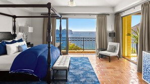 Un lit à baldaquin avec vue sur la mer, un balcon avec un fauteuil et une grande fenêtre avec des rideaux.