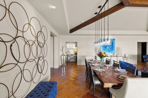 A modern dining area with a long wooden table set for six, surrounded by chairs. A blue upholstered bench is visible in the foreground. The room features a high ceiling with exposed wooden beams and a large abstract wall art piece. - Stay 4+ Nights & Save Up to 25% - Deluxe 4BR PH -Ocean Views in Cabo (Cabo San Lucas)