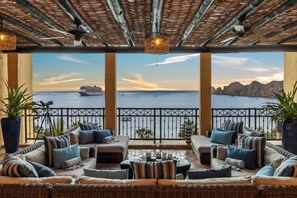 A balcony with wicker furniture, potted plants, and a view of the sea and mountains.