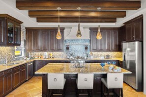 A modern kitchen with dark wood cabinets, a large island with bar stools, a stainless steel refrigerator, and pendant lighting. - Stay 4+ Nights & Save Up to 25% - Deluxe 4BR PH -Ocean Views in Cabo (Cabo San Lucas)