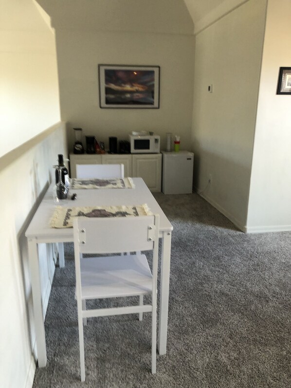 A small, clean room with a white table and chair, a microwave, a refrigerator, and a painting on the wall.