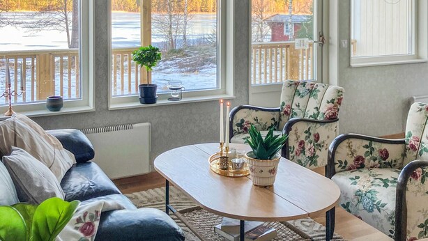 Ein gemütliches Wohnzimmer mit einem Holztisch, floralen Sesseln und Blick auf eine verschneite Landschaft durch große Fenster.