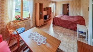 A room with a bed, a wooden table with a crocheted tablecloth, a wicker chair, a wardrobe, and a window with curtains.