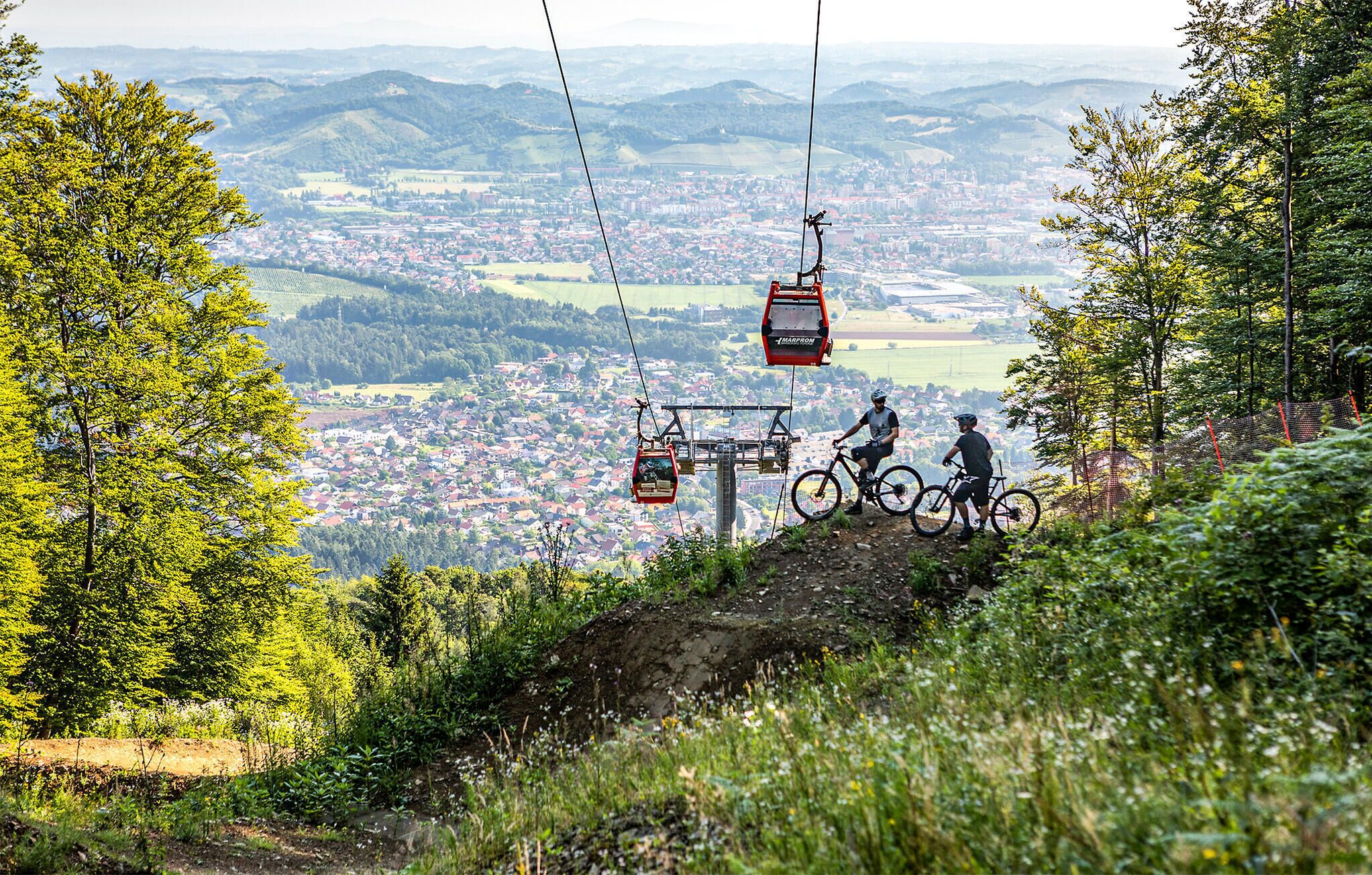 A mountain landscape with a ski lift, a town in the valley, and cyclists on a trail.