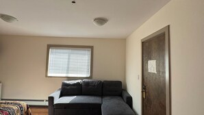 Ein Zimmer mit einer Couch, einem Bett, einem Fenster mit Jalousien, einer Tür und einer Decke mit Einbauleuchten.