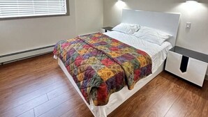Ein Schlafzimmer mit Holzboden, einem Bett mit bunter Steudecke, einem weißen Kopfteil und einem Nachttisch mit Schublade.